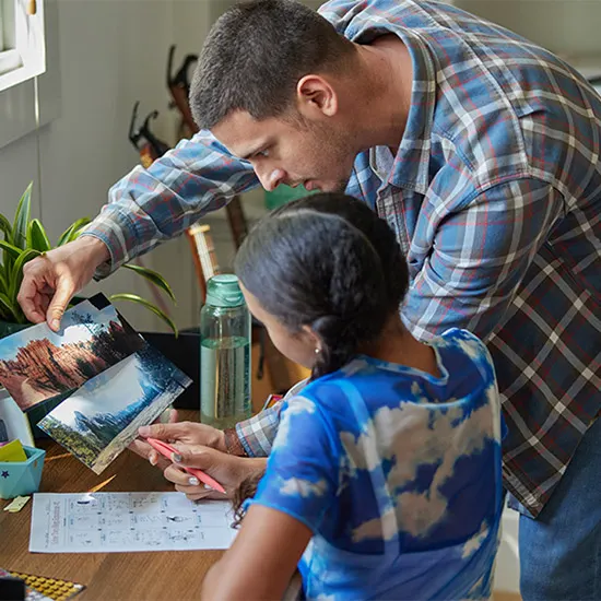 Ein Mann im karierten Hemd zeigt einem Kind Bilder auf einem Tisch mit Papier und einer Wasserflasche.