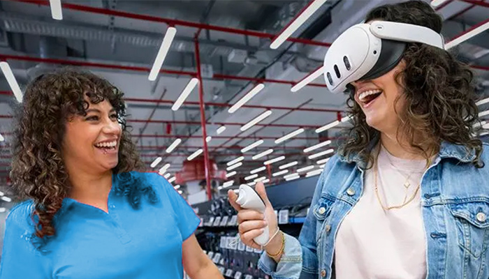 Zwei Frauen lachen in einem Geschäft. Eine trägt ein VR-Headset und hält einen Controller. Neonröhren sind an der Decke.