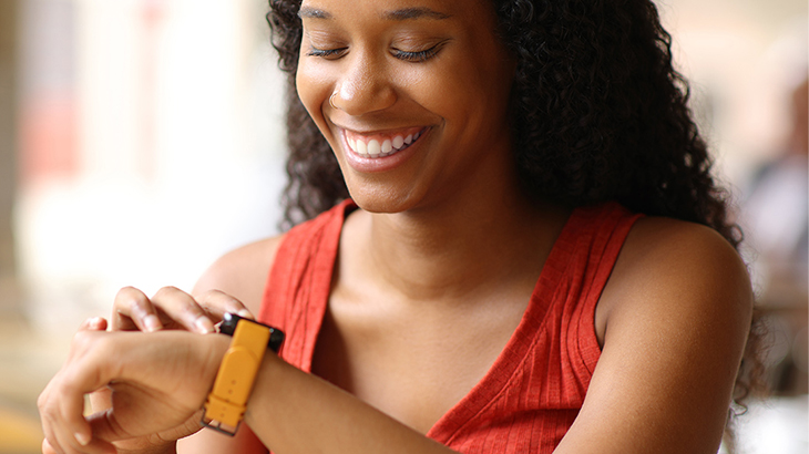 Eine lächelnde Person mit rotem Top schaut auf eine Smartwatch mit gelbem Armband am Handgelenk.