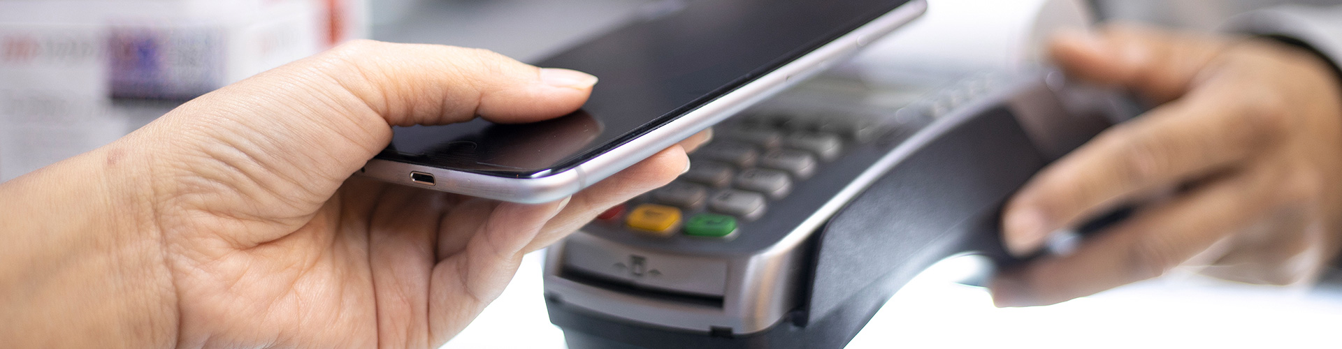 Eine Hand hält ein schwarzes Smartphone über ein graues Zahlungsgerät mit Tasten und einem kleinen Bildschirm.