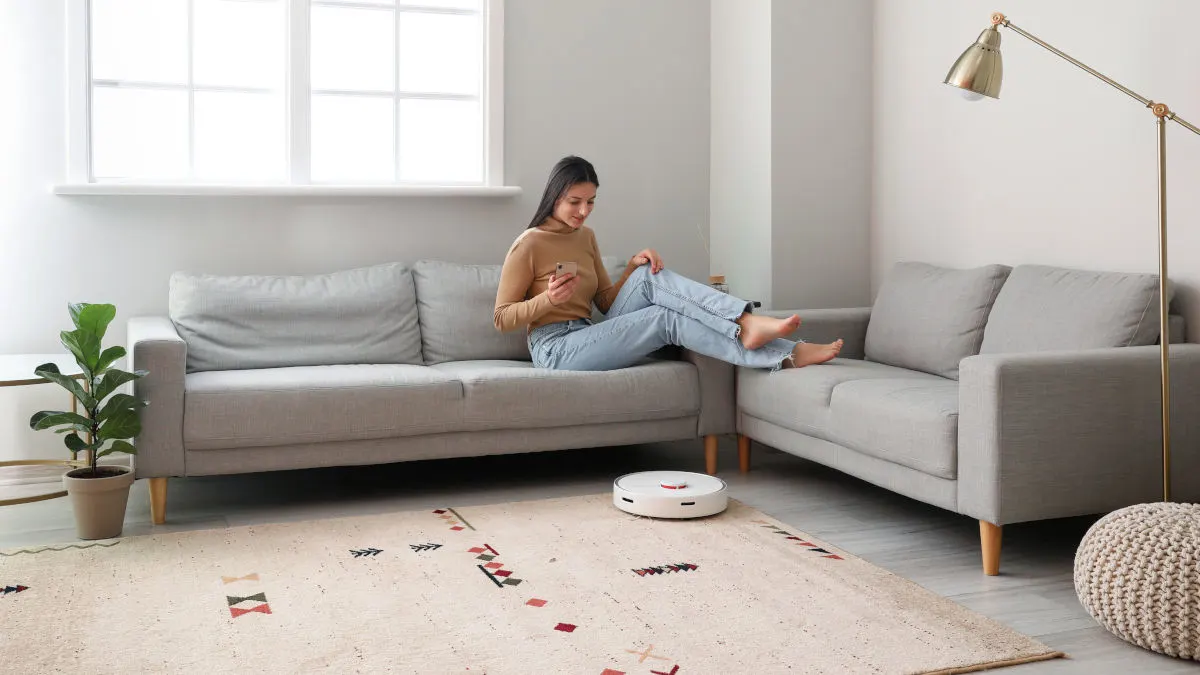 Eine Frau sitzt auf einem grauen Sofa und benutzt ihr Handy. Ein Staubsaugerroboter reinigt den Teppich im Wohnzimmer.