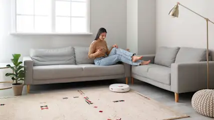 Eine Frau sitzt auf einem grauen Sofa und benutzt ihr Handy. Ein Staubsaugerroboter reinigt den Teppich im Wohnzimmer.