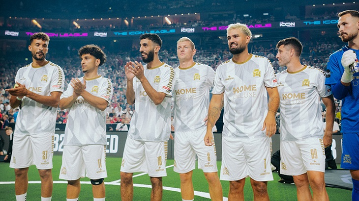 Eine Gruppe von Fußballspielern in weißen Trikots mit goldenen Akzenten steht auf einem grünen Feld vor Zuschauern.