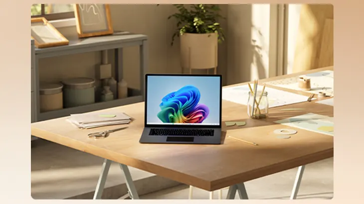 Auf einem Holztisch steht ein Laptop mit buntem Bildschirm. Stifte im Glas, Papier und eine Pflanze sind auch zu sehen.