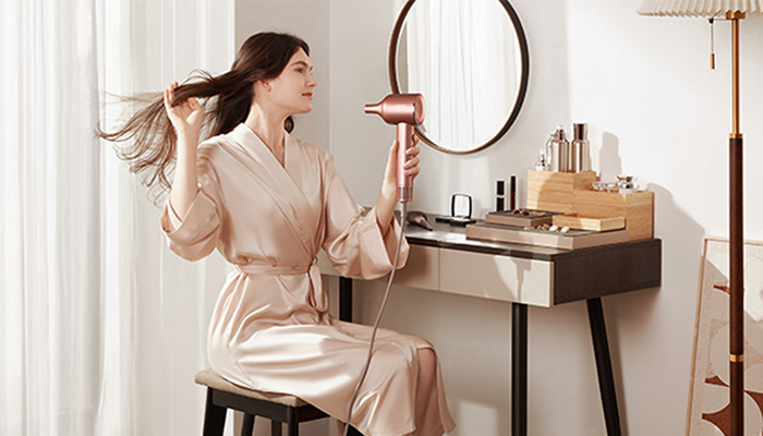 Eine Frau im Bademantel sitzt vor einem Schminktisch und föhnt ihre Haare, ein runder Spiegel hängt an der Wand.