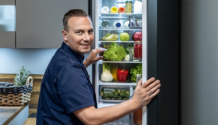 Ein Mann öffnet einen Kühlschrank gefüllt mit Gemüse, Obst und eingelegten Produkten. Eine Ananas steht auf einem Tisch.