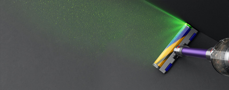 Ein Staubsauger mit blau-gelber Bürste und grünem Laserstrahl steht auf einem grauen Untergrund, der mit kleinen Partikeln bedeckt ist.