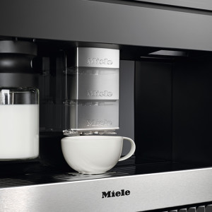 Nahaufnahme einer Miele Kaffeemaschine mit Milchbehälter und Tasse. Die Maschine ist schwarz und silber.