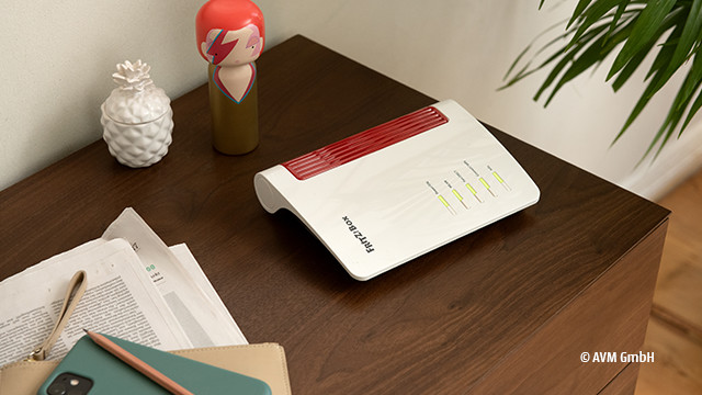 Ein Router liegt auf einem braunen Tisch, zusammen mit Papieren, Stiften und Deko-Objekten wie einer Ananas und einer Holzfigur.