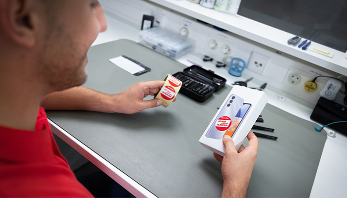 Eine Person in lila Jacke und orangefarbenem Pullover hält eine weiße Schachtel mit grünem Text. Im Hintergrund sind ein rotes Wandbild und Deckenleuchten zu sehen.