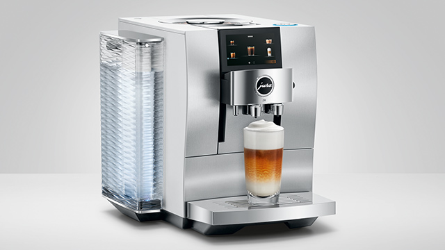 Silberne Kaffeemaschine mit Tassenbehälter, die ein Getränk in ein Glas ausgibt. Der Hintergrund ist hellgrau.