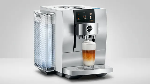 Silberne Kaffeemaschine mit Tassenbehälter, die ein Getränk in ein Glas ausgibt. Der Hintergrund ist hellgrau.