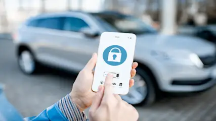 Eine Person hält ein Smartphone mit einer blauen Schloss-App vor einem silbernen Auto. Der Finger berührt den Bildschirm.
