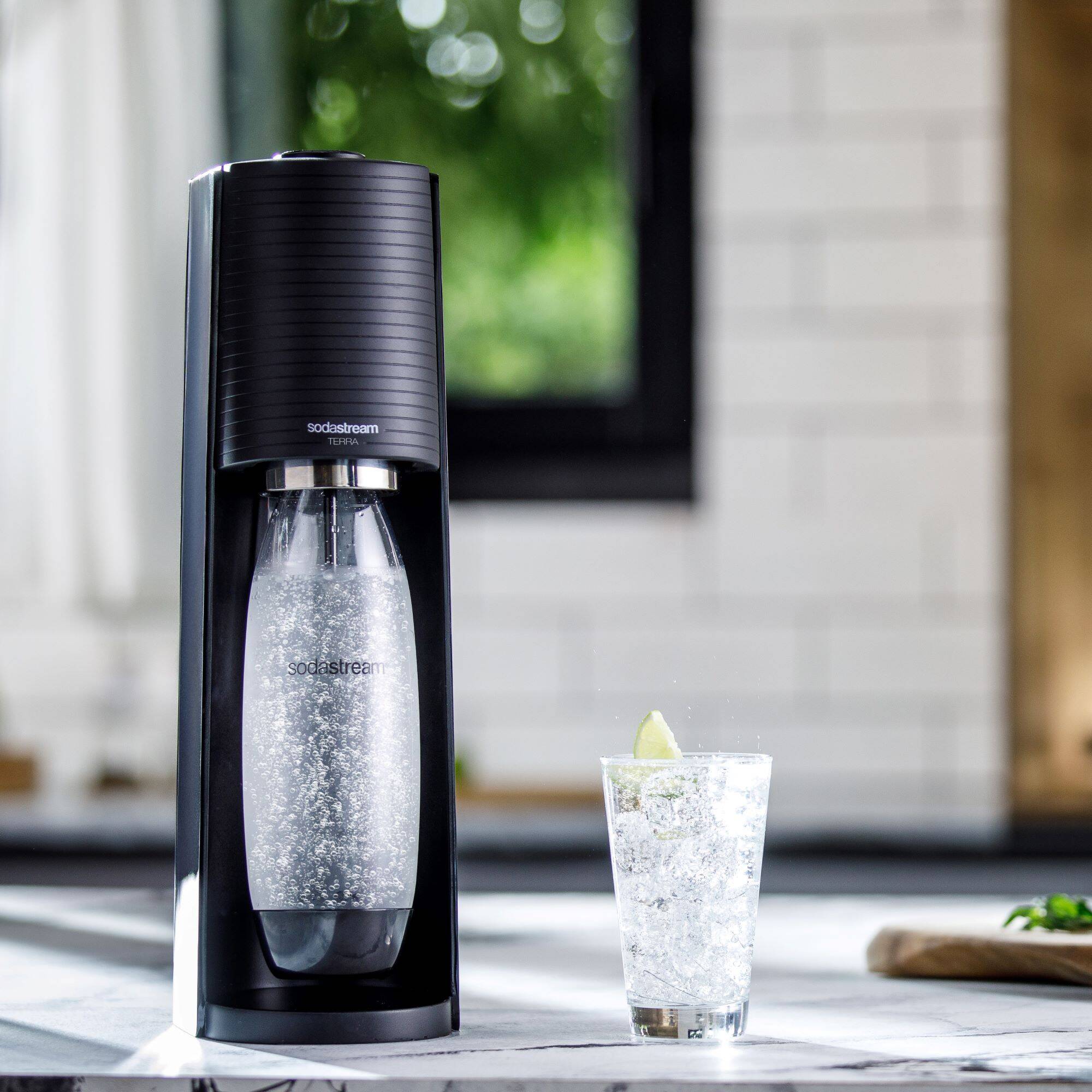 Ein SodaStream Wassersprudler in Schwarz steht neben einem Glas Sprudelwasser mit Limette auf einer Küchentheke.
