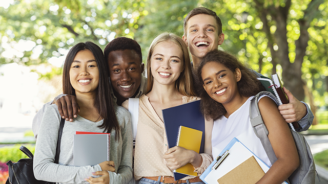 Eine Gruppe von fünf lächelnden Studenten steht draußen mit Büchern und Rucksäcken vor Bäumen.