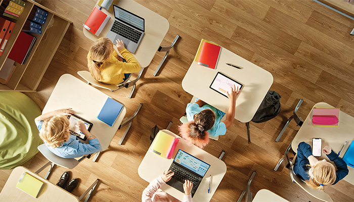 Blick von oben in eine Grundschulklasse. Schüler sitzen an separaten Schreibtischen, vor ihnen liegt jeweils ein Notebook oder ein Tablet.