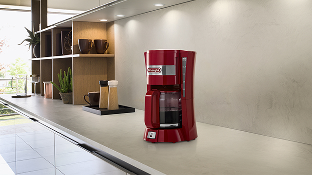 Eine rote Kaffeemaschine steht auf einer Küchentheke, neben Regalen mit Tassen und Pflanzen unter hellen Lichtern.