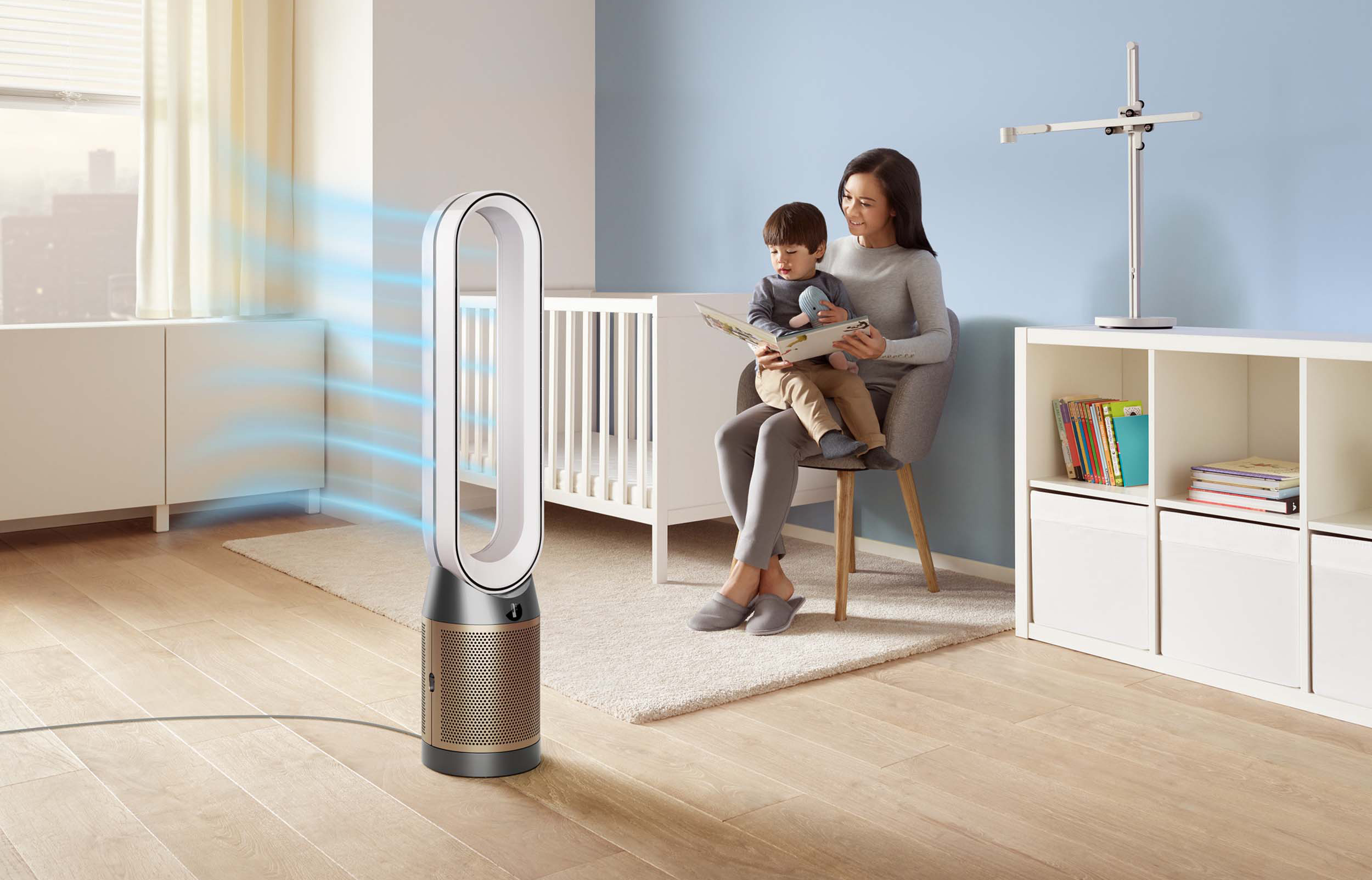 Mutter liest Kind ein Buch, sitzend auf einem Stuhl. Ein weißes Kinderbett und ein Ventilator stehen im Raum.