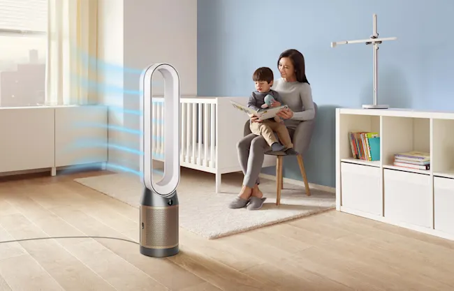 Mutter liest Kind ein Buch, sitzend auf einem Stuhl. Ein weißes Kinderbett und ein Ventilator stehen im Raum.