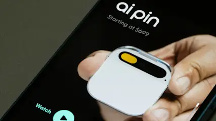 Nahaufnahme eines Telefons, das ein weißes, quadratisches Gerät mit gelbem Akzent und den Text "ai pin" anzeigt.