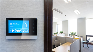 Ein digitales Thermostat an einer weißen Wand mit einem blauen Bildschirm neben einem Büroraum mit Schreibtischen und Stühlen.