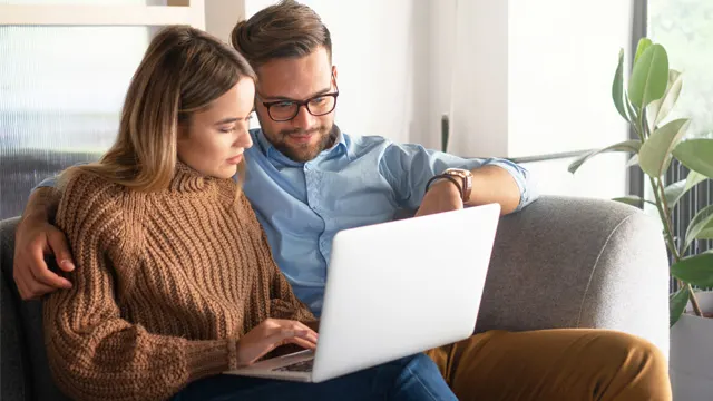 Ein Paar sitzt auf einem grauen Sofa und benutzt einen Laptop. Der Mann trägt ein blaues Hemd, die Frau einen braunen Pullover.