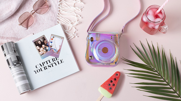 Auf einem rosafarbenen Tisch liegen eine Zeitschrift, eine Kamera, eine Sonnenbrille, ein Getränk, ein Eis und ein Palmblatt.