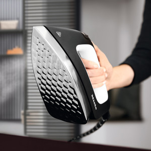 Person hält ein schwarzes und silbernes Bügeleisen mit weißem Griff vor einem Regal und Jalousien.