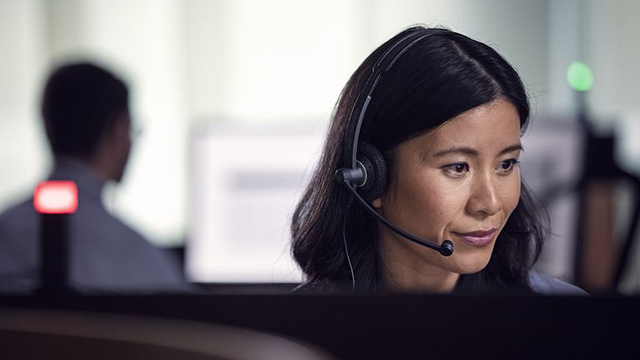 Eine Frau mit Headset arbeitet am Computer, ein Mann im Hintergrund ist verschwommen.
