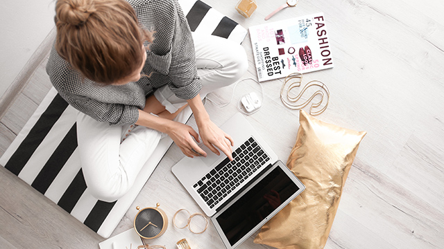 Eine Person sitzt auf einem gestreiften Kissen und tippt auf einem Laptop, umgeben von einem Magazin, einer Uhr und Accessoires.