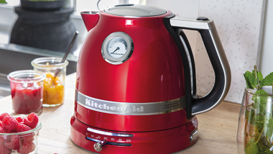 Ein roter KitchenAid-Wasserkocher mit Temperaturanzeige steht auf einer Küchentheke, neben Gläsern mit Früchten und Marmelade.