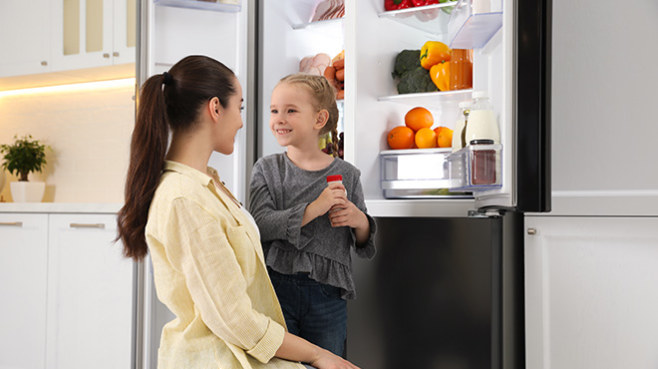 Eine Mutter hockt vor einem geöffneten Kühlschrank, ihre Tochter hält ein Getränk in der Hand.