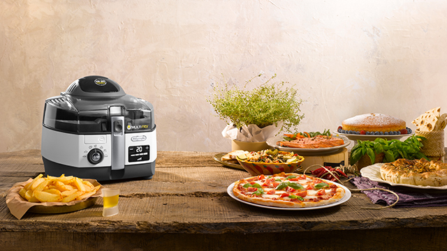 Auf einem Tisch stehen eine Fritteuse, Pommes, Pizza, Kuchen und ein Topf mit grünen Pflanzen.