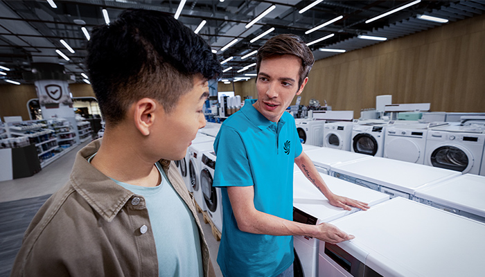 Ein Mann in blauem Hemd zeigt einem anderen Mann in einer Abteilung mit Haushaltsgeräten eine Waschmaschine.