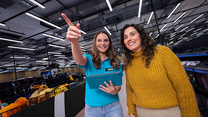 Zwei Frauen in einem Laden. Eine zeigt mit dem Finger nach oben, hält ein blaues Tablet und trägt ein blaues Hemd mit Logo.