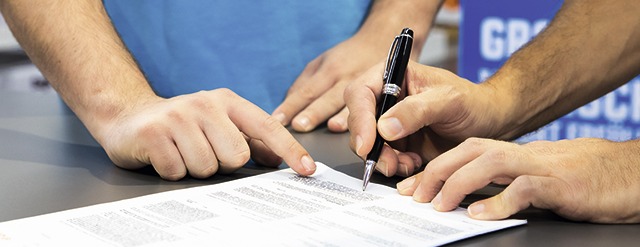 Ein Dokument wird mit einem schwarzen Stift unterschrieben, während eine Hand darauf zeigt, auf einem grauen Tisch.