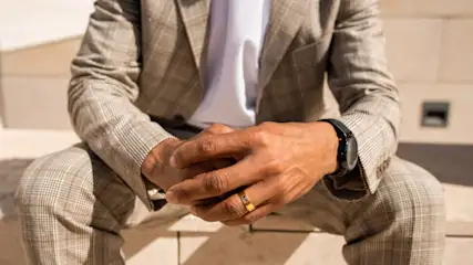 Ein Mann im karierten Anzug sitzt mit verschränkten Händen, trägt eine Uhr und einen Ring. Der Hintergrund ist eine Wand.