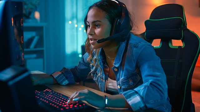 Eine Frau mit Headset tippt auf einer rot beleuchteten Tastatur, während sie vor einem Computer in einem Gaming-Stuhl sitzt.