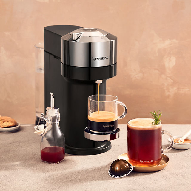 Eine Kaffeemaschine füllt eine Tasse Kaffee. Eine zweite Tasse Kaffee steht daneben, zusammen mit Keksen und einer Flasche Sirup.