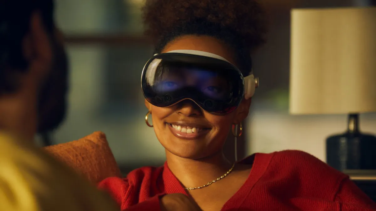 Eine Frau lächelt und trägt eine schwarze VR-Brille, goldene Ohrringe und eine goldene Halskette, mit einem roten Pullover.