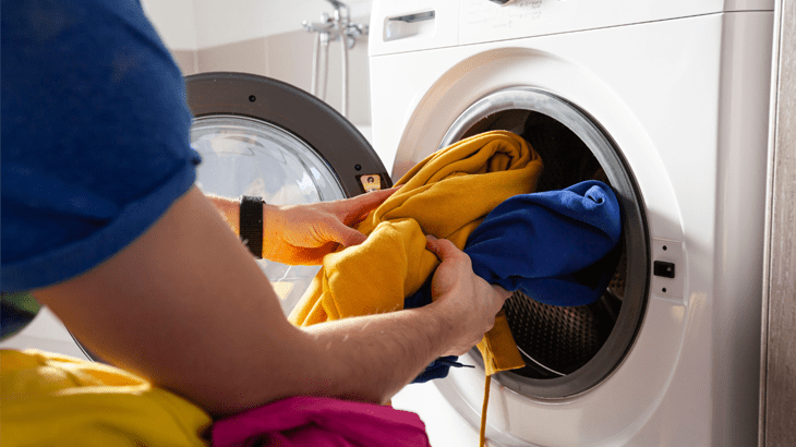 Eine Person mit blauem Hemd leert eine weiße Waschmaschine mit gelber und blauer Kleidung.