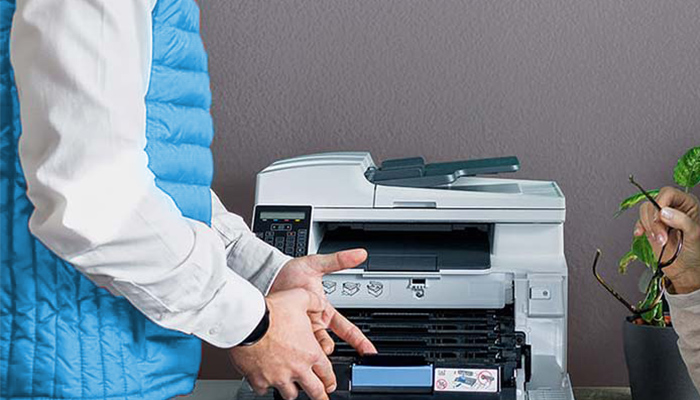 Eine Person in blauer Weste bedient einen Drucker vor einer grauen Wand. Eine Hand hält eine Brille neben einer Topfpflanze.