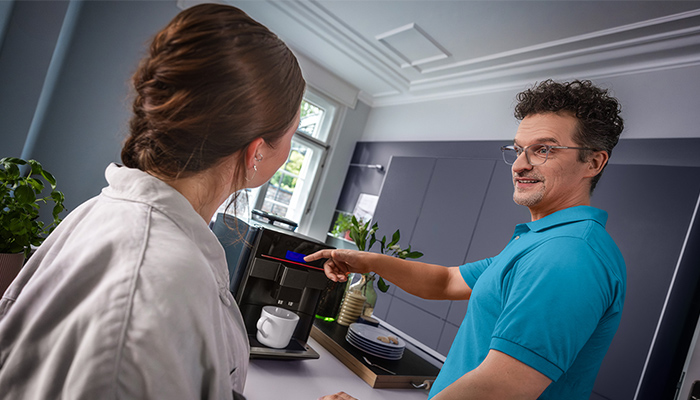 In der Küche zeigt ein Mann einer Frau eine Kaffeemaschine. Tasse, Teller und Pflanzen sind auch sichtbar.