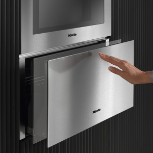 Edelstahl-Backofen mit ausziehbarer Schublade an einer dunklen Wand, eine Hand öffnet die Schublade.