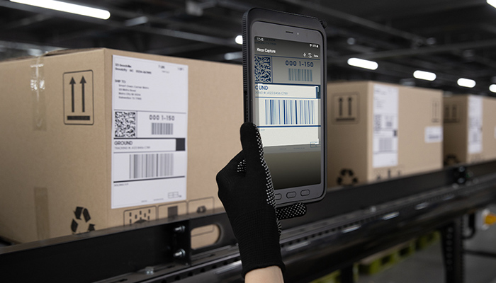 Eine Person scannt ein Paket, das ein Fließband entlang fährt, mit seinem Smartphone.