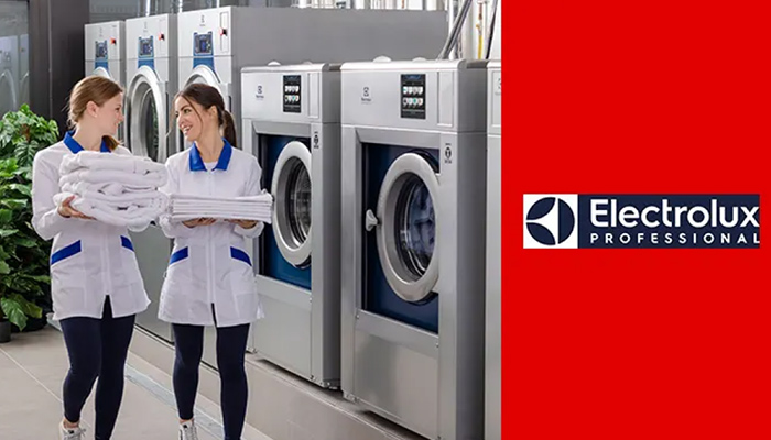 Zwei Frauen tragen je einen Stapel Wäsche, während sie an diversen Waschmaschinen vorbegehen.