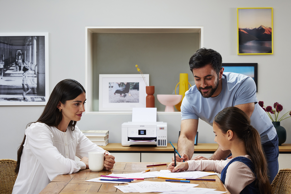 Eine Familie sitzt am Tisch, ein Drucker steht bereit. Die Tochter malt, während die Eltern zusehen und helfen.