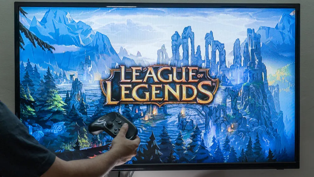 Ein Fernseher zeigt das Spiel "League of Legends" mit Bergen, Bäumen und Burgen im Hintergrund. Eine Person hält einen Controller.