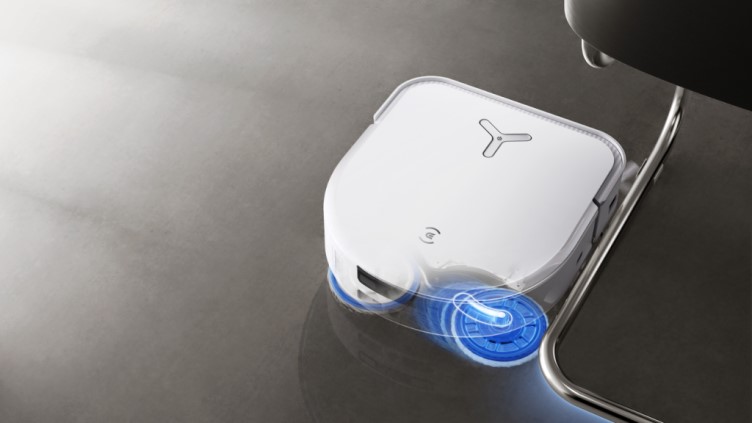 Ein weißer Roboter-Staubsauger mit blauen Rädern steht auf einem grauen Untergrund neben einem Stuhlbein aus Metall.