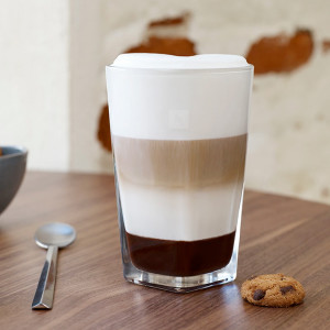 Ein Glas Latte Macchiato mit drei Schichten auf einem Tisch, daneben ein Löffel und ein Keks, vor einer Wand.